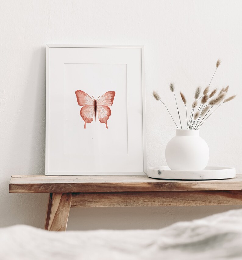 May include: A framed watercolor painting of a pink butterfly on a white background. The painting is displayed on a wooden shelf with a white vase of dried grasses.