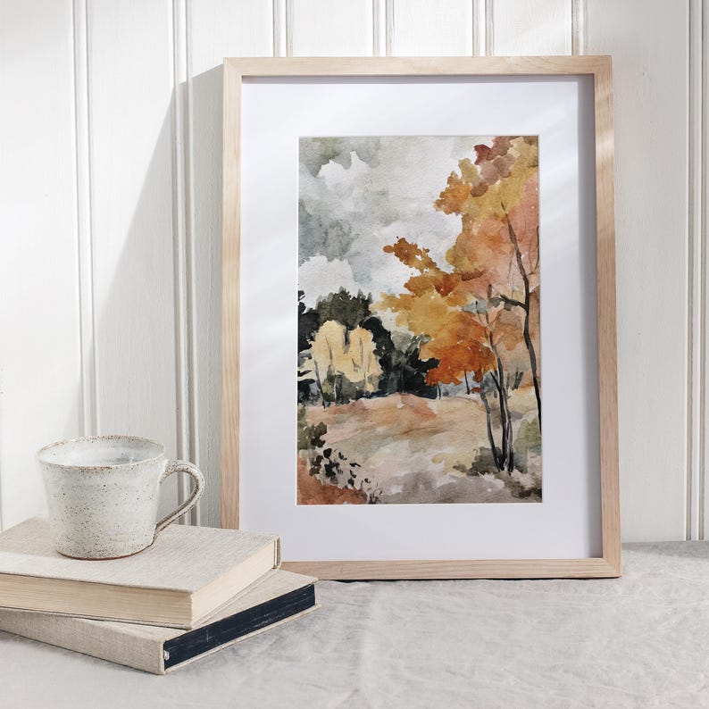 May include: Watercolor landscape art print in a light wood frame. The artwork depicts a fall scene with trees in shades of orange, yellow, and green. A ceramic mug and two books are in the foreground.
