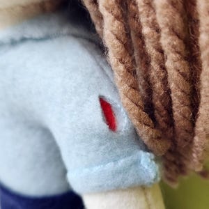 Pode incluir: Uma boneca de pel&uacute;cia azul claro com cabelo de l&atilde; marrom. A boneca tem um remendo vermelho no ombro.