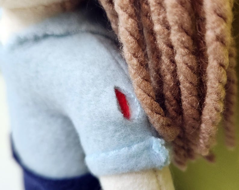 Pode incluir: Uma boneca de pel&uacute;cia azul claro com cabelo de l&atilde; marrom. A boneca tem um remendo vermelho no ombro.