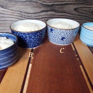 Könnte beinhalten: Vier Keramikschalen gefüllt mit einer weißen Substanz, möglicherweise Joghurt oder einem ähnlichen Lebensmittel. Die Schalen sind in Blautönen gehalten und weisen Muster wie Streifen, florale Designs und ein sternförmiges Motiv auf. Die Schalen sind auf einer Holzoberfläche angeordnet.