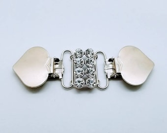 May include: A silver-toned metal clasp with a heart-shaped end on each side. The center features a row of eight clear, round rhinestones. The clasp is designed to fasten two pieces of fabric or material together.
