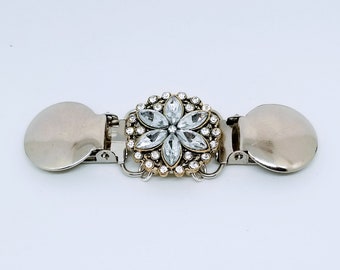 May include: Silver-toned metal brooch with a central floral design. The flower is adorned with clear, sparkling crystals and surrounded by smaller crystals. The brooch has two rounded, silver-colored clasps on either end.