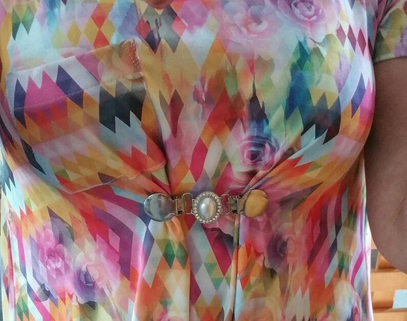 May include: A colorful patterned shirt with a silver and pearl brooch. The shirt has a geometric pattern with pink, yellow, blue, and green colors. The brooch is in the center of the shirt and has a pearl in the center.