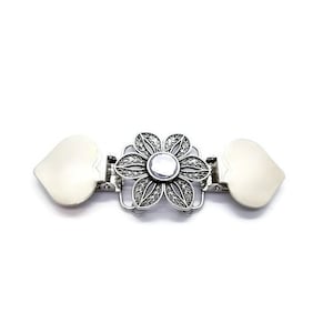K&ouml;nnte beinhalten: Ein silberfarbener Metallverschluss mit einem floralen Design in der Mitte und herzf&ouml;rmigen Enden. Die Blume hat einen zentralen Edelstein und detaillierte Bl&uuml;tenblattmuster. Der Verschluss dient zum Verbinden von Stoff- oder Materialteilen.