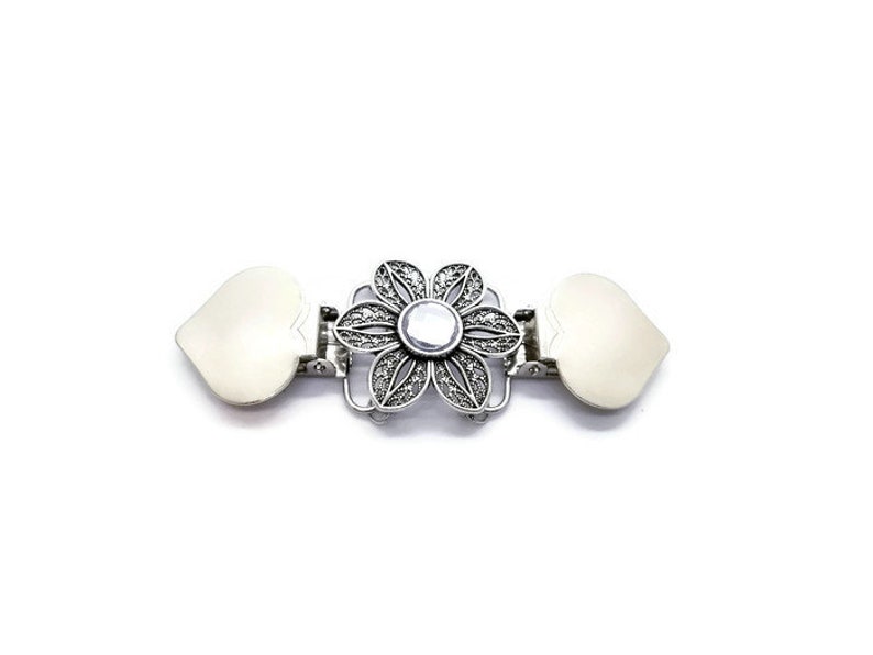 K&ouml;nnte beinhalten: Ein silberfarbener Metallverschluss mit einem floralen Design in der Mitte und herzf&ouml;rmigen Enden. Die Blume hat einen zentralen Edelstein und detaillierte Bl&uuml;tenblattmuster. Der Verschluss dient zum Verbinden von Stoff- oder Materialteilen.