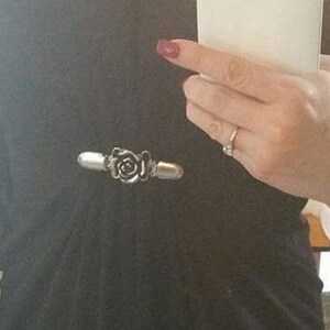 May include: A silver-toned rose-shaped brooch with a black center. The brooch is attached to a black shirt.