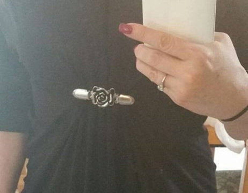May include: A silver-toned rose-shaped brooch with a black center. The brooch is attached to a black shirt.