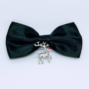 May include: A black bow tie with a silver reindeer charm. The reindeer has a red nose. The bow tie is made of a smooth, dark fabric. The reindeer charm is attached to the center of the bow tie.