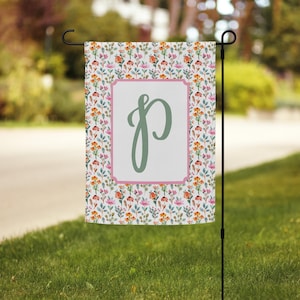 May include: A white garden flag with a pink and green floral pattern. The flag has a pink border and a green letter "P" in the centre.