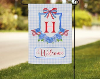 May include: A garden flag with a blue and white gingham pattern. The flag features a red letter "H" inside a blue and white shield, with a red and blue bow at the top. Two American flags flank the shield, and the word "Welcome" is written at the bottom.