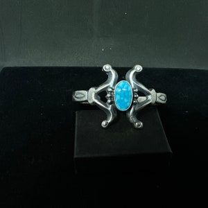 Puede incluir: Un brazalete de plata con una piedra turquesa azul engastada en el centro. El brazalete tiene un diseño único con dos brazos curvos que se unen en la piedra.