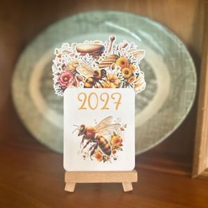 May include: A white square calendar card for the year 2027, featuring a detailed illustration of a honeybee surrounded by small yellow flowers. The card is topped with a decorative cutout of bees, honey dippers, and sunflowers, resting on a wooden stand.
