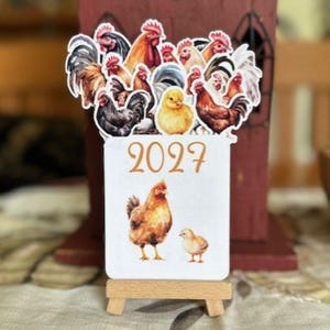 May include: A rectangular 2027 calendar card featuring a collection of colorful rooster and chick illustrations at the top, with a hen and a chick printed below the year. The card is displayed on a small, light-colored wooden easel stand.