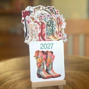 May include: A 2027 calendar card featuring a pair of blue cowboy boots adorned with orange and pink roses, displayed on a small wooden easel. Behind the card is a layered cutout illustration of various cowboy boots with floral patterns.