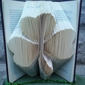 May include: A book folded into the shape of a three-leaf clover. The book is open and the pages are folded to create a three-dimensional shape. The book is on a green surface.