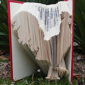 May include: A book art sculpture of a rooster, made by folding the pages of a book. The book is open and the rooster is standing on the pages. The book has a red cover.