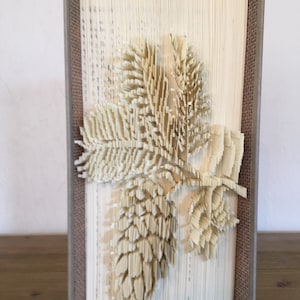Puede incluir: Una escultura de arte de libro de una piña y hojas hecha de las páginas de un libro. El libro está encuadernado en tela marrón y las páginas están dobladas en un diseño 3D.