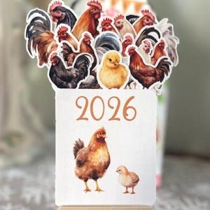 May include: A small wooden easel displays a calendar for the year 2026, featuring watercolor illustrations of chickens and a chick. Above, various rooster and hen stickers are arranged. The background includes a decorative element.