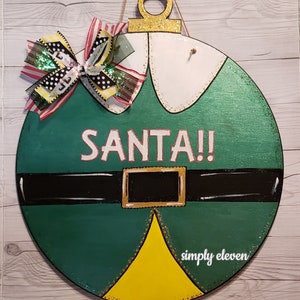 May include: A green and yellow Christmas ornament with a white collar and black belt. The ornament has the word "SANTA!" written on it in white letters. The ornament is decorated with gold glitter and a red, white, and green bow.