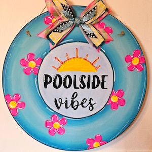 May include: A blue round wooden door hanger with a yellow sun, pink flowers, and the text "Poolside vibes". A pink and white bow with black polka dots is attached to the top.