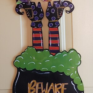 May include: A black cauldron with green bubbling liquid and a sign that reads "BEWARE". The cauldron is decorated with a witch's legs and feet, and a green, purple, and orange bow is hanging above it. The sign also includes the text "simply eleven".