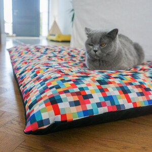 May include: A gray cat with yellow eyes is lying on a colorful rectangular pet bed with a black border. The bed has a pixelated pattern with squares of various colors, including red, blue, green, yellow, and white.