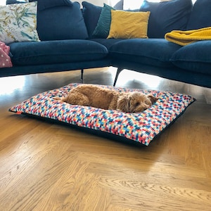 May include: A brown dog sleeping on a colorful patterned dog bed with a black border. The bed is on a wooden floor.
