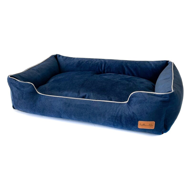 K&ouml;nnte beinhalten: Ein marineblaues, rechteckiges Haustierbett mit samtiger Textur. Das Bett hat eine wei&szlig;e Paspel an den R&auml;ndern und ein braunes Lederetikett. Dieses bequeme Bett ist f&uuml;r kleine bis mittelgro&szlig;e Hunde konzipiert und bietet einen gem&uuml;tlichen Ruheplatz.