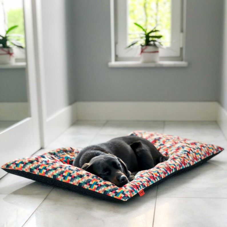 May include: A black dog is sleeping on a colorful rectangular dog bed with a black border. The bed is on a white tiled floor.