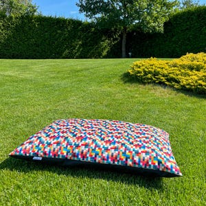 May include: A colorful rectangular dog bed with a geometric pattern of squares in shades of red, orange, yellow, green, blue, and white. The bed has a black border and is lying on a green grassy lawn.