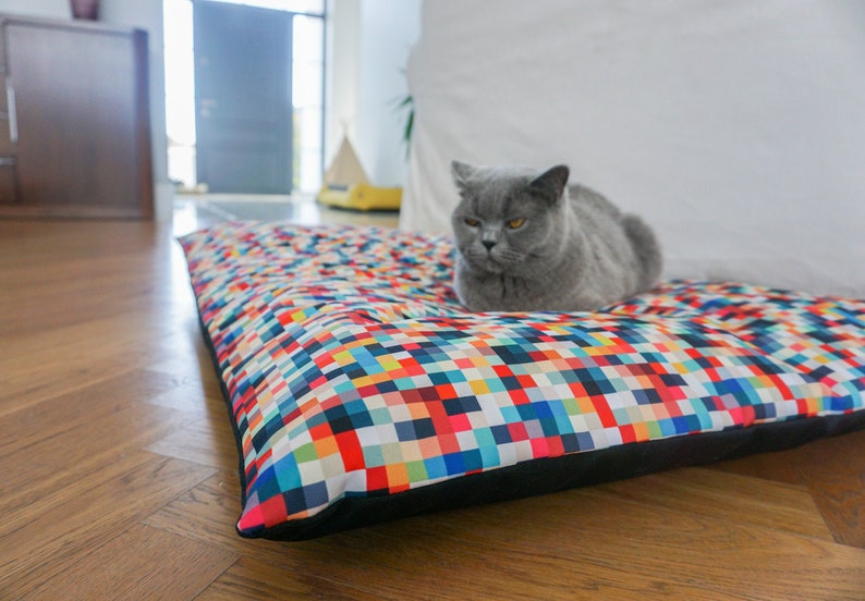 May include: A gray cat with yellow eyes is lying on a colorful rectangular pet bed with a black border. The bed has a pixelated pattern with squares of various colors, including red, blue, green, yellow, and white.