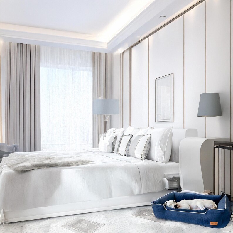 K&ouml;nnte beinhalten: Ein luxuri&ouml;ses Schlafzimmer mit einem gro&szlig;en wei&szlig;en Bett, weichen Kissen und einer grauen Decke. Ein marineblaues Hundebett steht auf dem Boden, in dem ein kleiner wei&szlig;er Hund schl&auml;ft. Der Raum hat wei&szlig;e W&auml;nde und ein gro&szlig;es Fenster.