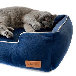 K&ouml;nnte beinhalten: Ein marineblaues Haustierbett mit cremefarbenem Rand und einem braunen Kunstlederetikett mit der Aufschrift "halka.life". Eine graue Katze liegt in dem Bett, die Pfoten verdecken ihr Gesicht. Das Bett ist aus weichem, pl&uuml;schigem Material.