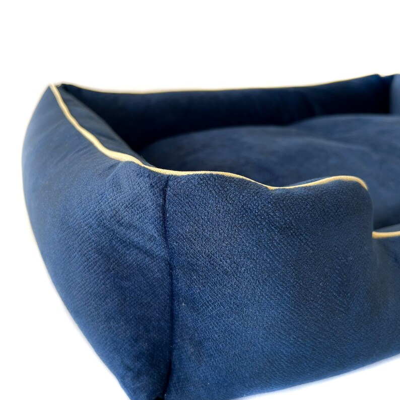 K&ouml;nnte beinhalten: Ein marineblaues Haustierbett mit strukturiertem Stoff und einer kontrastierenden goldfarbenen Paspel. Das Bett hat eine gepolsterte Innenseite und erh&ouml;hte Seiten f&uuml;r Komfort. Das Design ist schlicht und elegant.