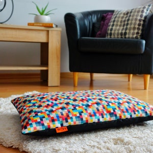 May include: A colorful rectangular dog bed with a geometric pattern. The bed is made of a soft, durable fabric and has a black border. The bed is sitting on a white fluffy rug.
