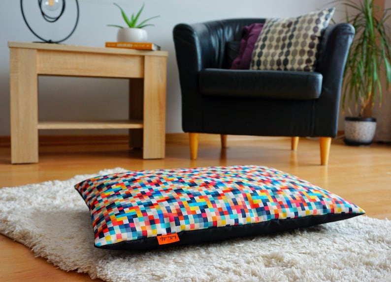 May include: A colorful rectangular dog bed with a geometric pattern. The bed is made of a soft, durable fabric and has a black border. The bed is sitting on a white fluffy rug.