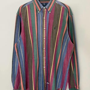 May include: A long-sleeve button-down shirt featuring a vertical striped pattern in shades of maroon, green, blue, and yellow. The shirt has a button-down collar, a front button placket, and a small green embroidered logo on the left chest area.