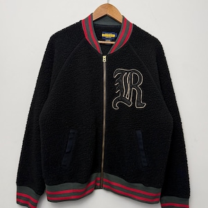 May include: A black sherpa fleece bomber jacket featuring a gold-tone front zipper, two side pockets, and a large embroidered letter R on the chest. The collar, cuffs, and hem are ribbed with grey and red stripes. A Rugby label is at the neck.