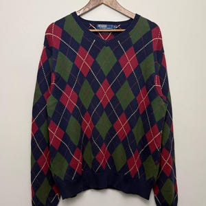 May include: A long-sleeve V-neck sweater featuring a multi-colored argyle pattern in navy blue, forest green, and deep red with thin white diagonal lines. The knit garment has a ribbed collar, cuffs, and hem, and includes a Polo by Ralph Lauren label.