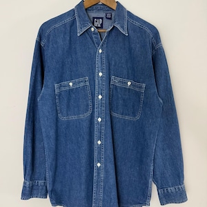 May include: A long-sleeved blue denim button-down shirt featuring a classic collar, two chest pockets with button closures, and white buttons down the front. The interior neck label displays the brand name GAP in white text on a dark blue background.