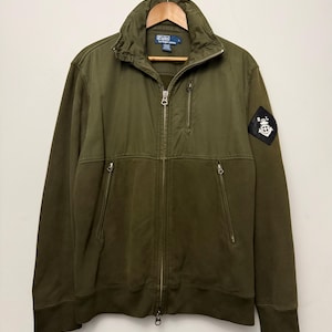 May include: An olive green zip-up jacket featuring a stand-up collar, a vertical zippered chest pocket, and two lower zippered pockets. A black diamond-shaped patch with a white anchor emblem and the letters RL is on the left sleeve.