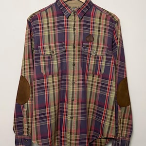 May include: A long-sleeve button-down shirt featuring a plaid pattern in shades of purple, red, and olive green. The shirt includes two chest pockets, a small circular brown patch on the left chest, and brown suede-like elbow patches on each sleeve.