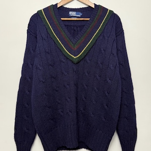 May include: A navy blue cable-knit sweater featuring a V-neckline with green, red, and yellow striped trim. The garment includes a Polo by Ralph Lauren label at the inner collar and long sleeves with ribbed cuffs and a ribbed hemline.