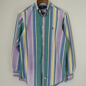 May include: A button-down shirt with vertical stripes in shades of green, purple, yellow, and blue. The shirt has long sleeves and a button-down collar. A small logo is visible on the chest.