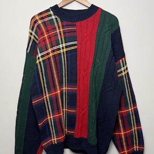 Pode incluir: Uma camisola de manga comprida com gola redonda, apresentando um design de retalhos com painéis vermelhos, verdes e azul-marinho. A peça combina secções com padrões de xadrez tartan e texturas de malha trançada, com uma gola canelada azul-marinho.