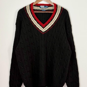 May include: Black cable-knit sweater with a V-neck. The neckline features a red, beige, and white striped design. The sweater is hanging on a wooden hanger. The label reads "Polo by Ralph Lauren".