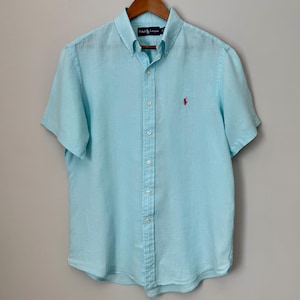 Puede incluir: Camisa azul claro de manga corta con un pequeño logo rosa en el pecho. La camisa está colgada de una percha de madera. La camisa tiene cuello abotonado y un corte clásico.