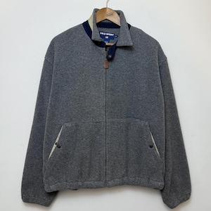 May include: A grey fleece zip-up jacket featuring a high collar with navy blue and beige accents. The garment includes a front zipper with a brown leather pull tab, two side pockets with snap closures, and a Polo Sport label at the neckline.