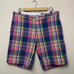 May include: A pair of plaid shorts featuring a vibrant pattern of blue, pink, green, yellow, and white intersecting lines. The shorts include a waistband with belt loops, a single white button closure, and a zipper fly, all hanging on a wooden hanger.